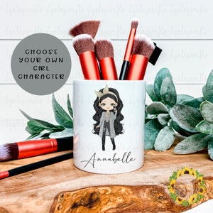 May include: White ceramic makeup brush holder with a cartoon girl illustration and the name "Annabelle" printed on the front. The holder is filled with makeup brushes.