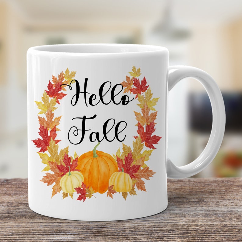 May include: White ceramic mug with a fall-themed design. The mug features a wreath of autumn leaves in shades of red, orange, and yellow, with the words "Hello Fall" in black script. Three pumpkins are centered in the wreath, one large orange pumpkin and two smaller white pumpkins.