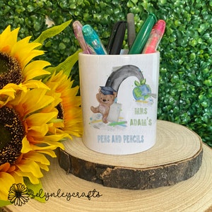 May include: White ceramic pencil holder with a rainbow, a teddy bear wearing a graduation cap, and the text "Mrs. Adam's Pens and Pencils".
