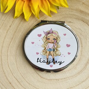 May include: A round compact mirror with a white background featuring a cartoon girl with blonde hair wearing a pink headband and a pink shirt. The girl is holding a phone and there are pink hearts scattered around her. The mirror has a silver frame and the name "Hayley" is written in black at the bottom.
