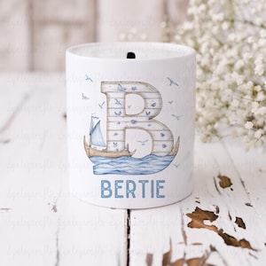 May include: White ceramic money box with a blue and brown nautical design. The letter "B" is featured on the front with a sailboat and waves. The name "BERTIE" is written below the design.