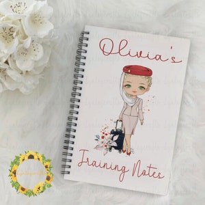 May include: A white spiral bound notebook with a cartoon illustration of a flight attendant holding a suitcase. The notebook has the text "Olivia's Training Notes" on the front.