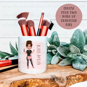 May include: White ceramic makeup brush holder with a pink and white design featuring a cartoon illustration of a woman with brown hair and a makeup brush. The text "KAT-MUA" is written vertically on the side of the holder.
