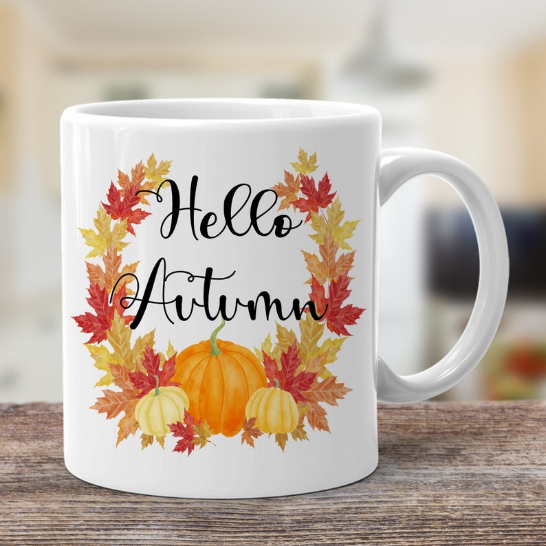 May include: White ceramic mug with a fall-themed design. The mug features a wreath of orange, red, and yellow leaves with the text "Hello Autumn" and three pumpkins in the center.