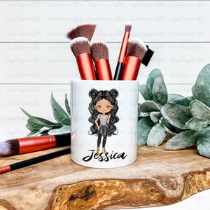 May include: White ceramic makeup brush holder with a cartoon illustration of a girl with black hair and brown eyes. The girl is wearing a black shirt, black pants, and brown boots. The text "Jessica" is printed on the holder.