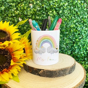 May include: White ceramic pencil holder with a rainbow, clouds, and flowers design. The holder has the text "Ms. Fox" written on the front.