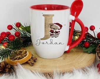 May include: White ceramic mug with a red interior and handle, featuring a gold letter "J" and a teddy bear in a Santa outfit. The mug includes a matching red spoon and the name "Jordan" in script. Festive holiday decor surrounds the mug.