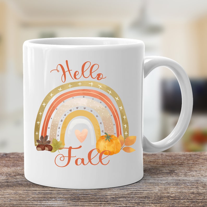 Może przedstawiać: Biały ceramiczny kubek do kawy z wzorem tęczy i tekstem "Hello Fall". Tęcza składa się z pomarańczowych, ż&oacute;łtych i beżowych pask&oacute;w z złotymi akcentami. Małe serce i dynia znajdują się w środku tęczy.