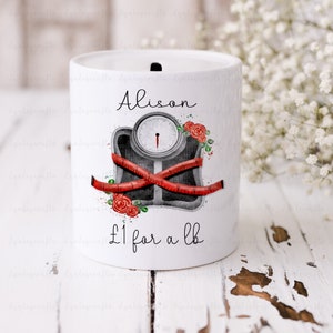 May include: White ceramic money jar with a personalized name "Alison" and a graphic of a scale with a red measuring tape wrapped around it. The graphic also includes red roses and the text "£1 for a lb".