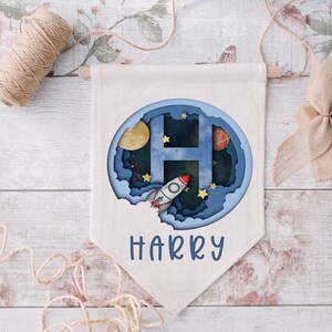 May include: A white canvas banner with a blue, paper-cut design of a rocket flying through space with stars, a moon, and a planet. The letter H is cut out of the design and the name HARRY is printed below.