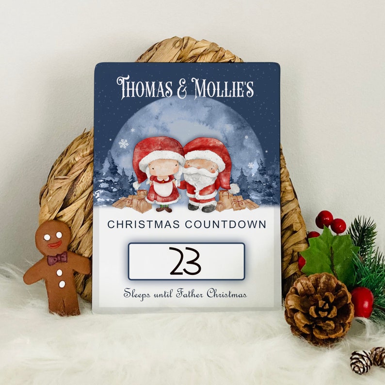 K&ouml;nnte beinhalten: Ein blaues und wei&szlig;es Weihnachts-Countdown-Schild mit den Namen "Thomas & Mollie's" oben. Das Schild zeigt eine Cartoon-Illustration von Weihnachtsmann und Frau Weihnachtsmann vor einer verschneiten Waldszene. Die Zahl "23" wird in einem wei&szlig;en Feld mit dem Text "Schl&auml;ft bis zum Weihnachtsmann" darunter angezeigt.