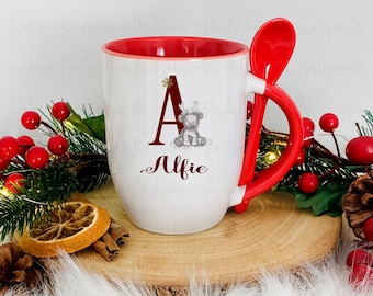 May include: White ceramic mug with a red rim and handle. The mug features a red spoon attached to the handle. The mug has a Christmas-themed design with a gray reindeer and the letter A. The mug is personalized with the name "Alfie".