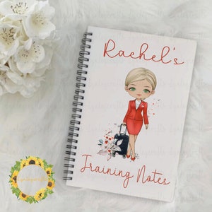 May include: A white spiral bound notebook with a cartoon illustration of a woman in a red uniform holding a suitcase. The notebook has the text "Rachel's Training Notes" printed on the front.