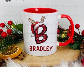 May include: White ceramic mug with a red rim and handle. The mug features a red glitter letter "B" with a Santa hat and reindeer antlers. The name "BRADLEY" is printed in red below the letter.