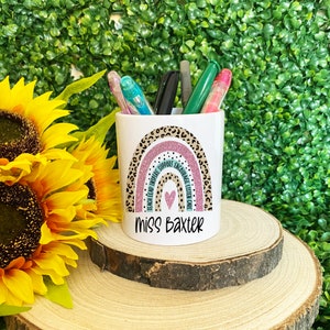 May include: White ceramic pencil holder with a rainbow design featuring a pink and teal color scheme. The rainbow has a leopard print pattern and the words "Teach Lead Inspire Support Encourage Listen Care" written around it. The holder has the name "Miss Baxter" written on the bottom.