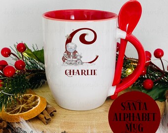 May include: White ceramic mug with a red rim and handle. The mug features a Santa Claus illustration with the letter "C" and the name "Charlie" printed on the front. The mug has a red spoon attached to the handle. The mug is on a wooden surface with a red circle that says "Santa Alphabet Mug".