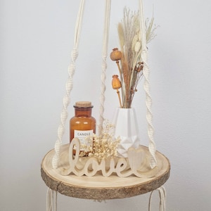 Könnte beinhalten: Runde Holz-Makramee-Ablage mit natürlicher Kante, mit einem braunen Glas-Kerzenglas, einer weißen geometrischen Vase mit Trockenblumen und einem Holzschild "love". Die Ablage wird von geflochtenen Seilen gehalten.