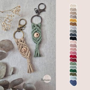 Könnte beinhalten: Zwei Makramee-Schlüsselanhänger in Beige und Salbeigrün, jeweils mit einem Holzknopf mit Pfotenabdruck. Die Schlüsselanhänger haben Metallringe und -verschlüsse. Auf der rechten Seite ist eine Farbkarte mit Makramee-Kordeloptionen abgebildet.