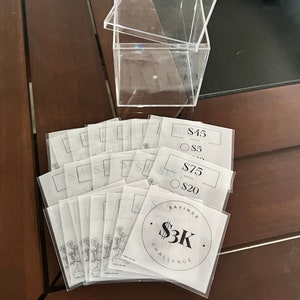 May include: A clear acrylic box with a lid, sitting on a wooden surface.  The box is filled with white paper slips with black text.  The slips have different amounts of money written on them, such as $5, $20, $45, and $75.  One slip has a circle with the text "Savings $3K Challenge".