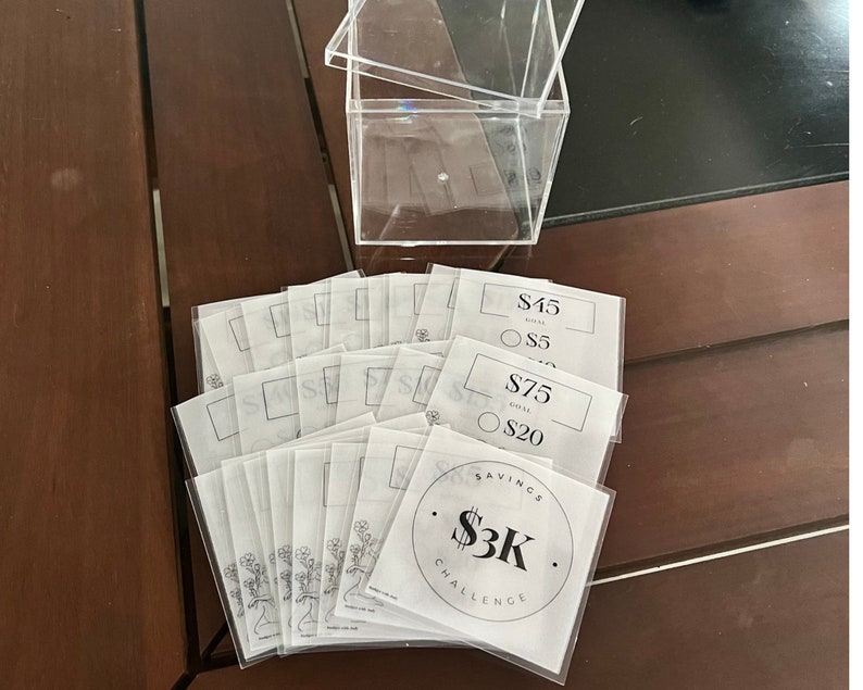 May include: A clear acrylic box with a lid, sitting on a wooden surface.  The box is filled with white paper slips with black text.  The slips have different amounts of money written on them, such as $5, $20, $45, and $75.  One slip has a circle with the text "Savings $3K Challenge".