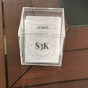 May include: A clear plastic box filled with white cards. The cards have black text on them, including the number "$105" and the text "$3K CHALLENGE".