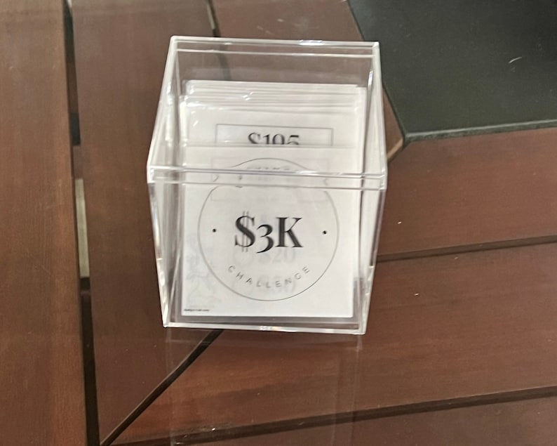 May include: A clear plastic box filled with white cards. The cards have black text on them, including the number "$105" and the text "$3K CHALLENGE".