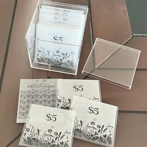 Pu&ograve; includere: Una scatola di acrilico trasparente piena di buste bianche con testo nero. Le buste sono etichettate con importi in dollari, tra cui $ 5, $ 30- $ 50, $ 55- $ 75 e $ 80- $ 100. La scatola &egrave; appoggiata su un tavolo di legno. Ci sono tre buste aggiuntive con un design floreale e il testo "$ 5" su un tavolo di legno.