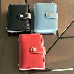 May include: Three leather card holders in different colors: black, red, and blue. Each holder has a single snap closure and a stitched edge.