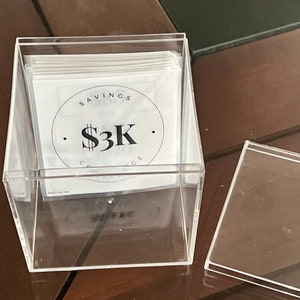 May include: A clear acrylic box filled with white cards. The top card has a black circle with the text "SAVINGS CHALLENGE" and "$3K" inside.