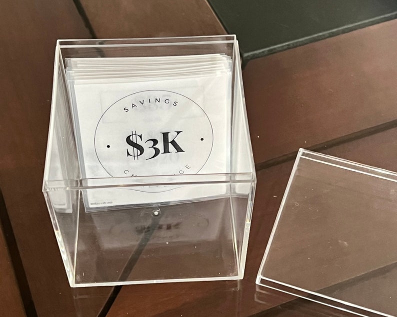 May include: A clear acrylic box filled with white cards. The top card has a black circle with the text "SAVINGS CHALLENGE" and "$3K" inside.