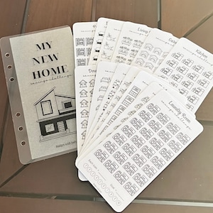 May include: A set of white sticker sheets with black outlines of various household items, such as a bed, couch, kitchen sink, and washing machine. The sticker sheets are labeled with the names of the rooms they represent, such as "Bedroom", "Living Room", "Kitchen", and "Laundry Room". The sticker sheets are arranged in a fan shape on a wooden surface. The text "My New Home Savings Challenge" is printed on a clear plastic sheet.