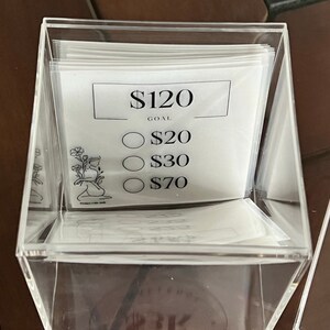 May include: A clear acrylic box with a white card inside. The card has a black border and the text "$120 GOAL" at the top. Below the text are three circles with the text "$20", "$30", and "$70" inside each circle.