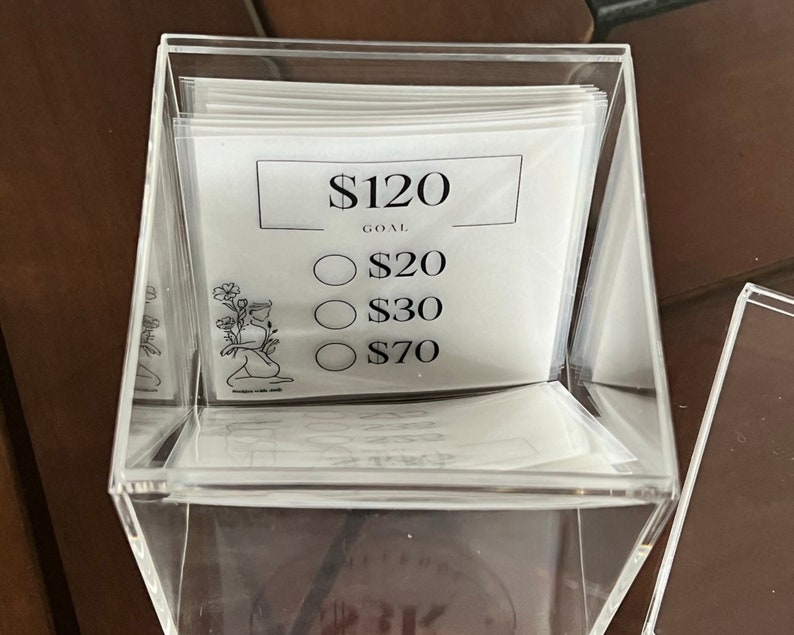 May include: A clear acrylic box with a white card inside. The card has a black border and the text "$120 GOAL" at the top. Below the text are three circles with the text "$20", "$30", and "$70" inside each circle.
