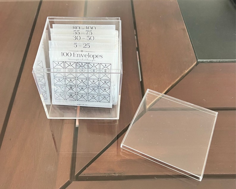 Pu&ograve; includere: Una scatola di plastica trasparente piena di 100 buste bianche. Le buste sono numerate da 5 a 100. La scatola ha un coperchio trasparente e il testo "100 Buste" &egrave; stampato sul davanti.