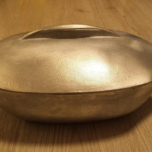 May include: A silver oval-shaped baking dish with a lid and a handle. The dish is made of metal and has a smooth surface.
