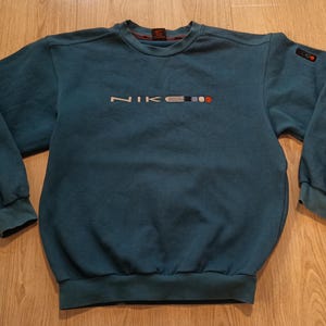 以下が含まれることがあります： 胸に白で「NIKE」のブランド名が刺繍された、ティール色のスウェットシャツ。ロゴには赤と青のアクセントがあります。長袖トップスはクルーネックとリブ編みの袖口が付いています。