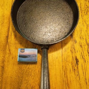 Puede incluir: Una sartén de hierro fundido negro con un mango largo, sobre una superficie de madera. Una pequeña caja de cerillas con una imagen de paisaje está al lado de la sartén. La sartén es redonda y tiene una superficie de cocción texturizada.