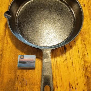 May include: A black cast iron skillet with a long handle. The skillet is empty and sitting on a wooden surface.