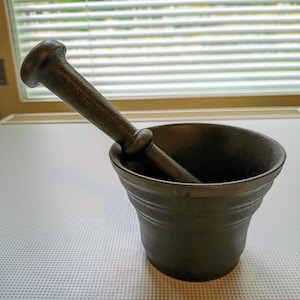 Könnte beinhalten: Ein schwarzer Mörser und Stößel aus Gusseisen. Der Mörser ist rund und hat eine strukturierte Oberfläche. Der Stößel ist lang und hat ein abgerundetes Ende.