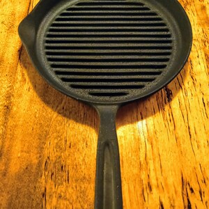 May include: A black cast iron grill pan with a long handle. The pan has a ridged surface for grilling.