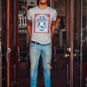 Peut inclure: Un homme portant un t-shirt gris avec un motif de canette de bi&egrave;re de style vintage qui dit "Buttweiser" et un jean bleu. Le motif de la canette pr&eacute;sente un sch&eacute;ma de couleurs rouge, blanc et bleu.