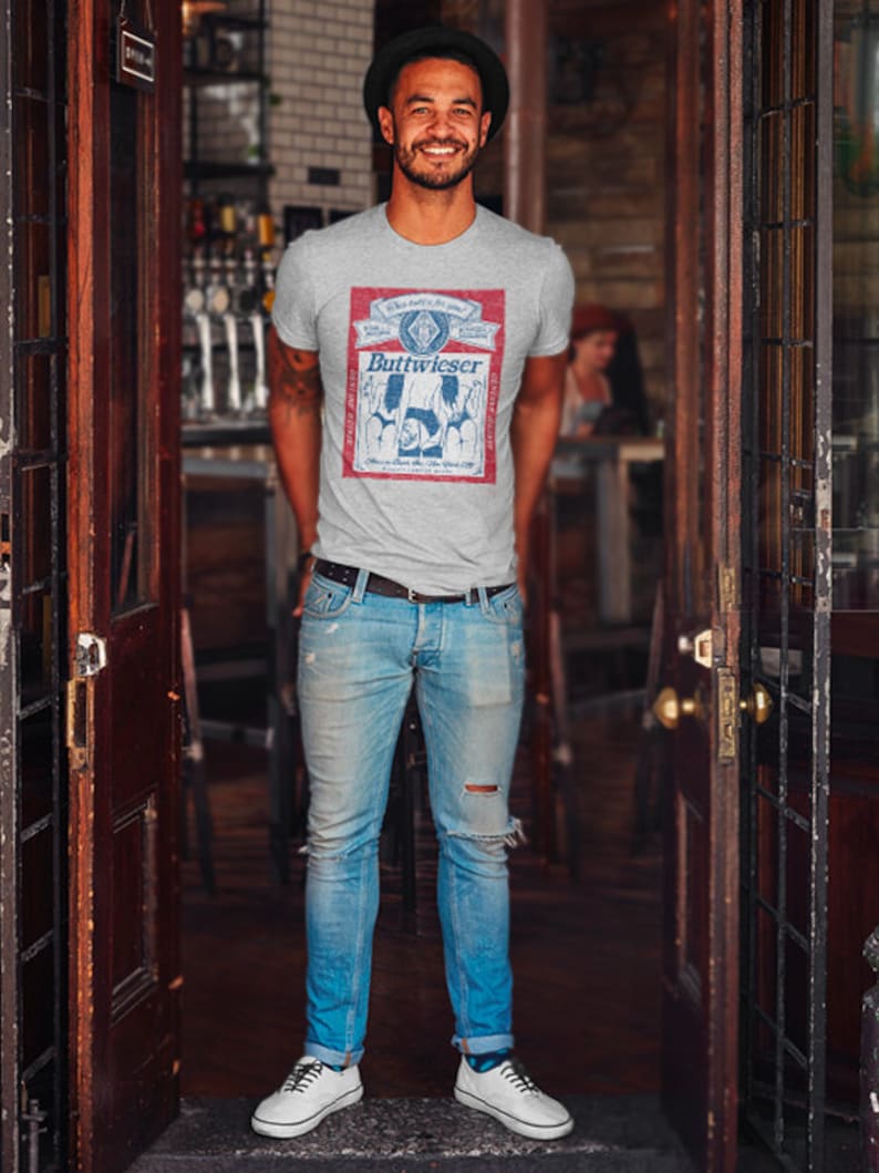 Peut inclure: Un homme portant un t-shirt gris avec un motif de canette de bi&egrave;re de style vintage qui dit "Buttweiser" et un jean bleu. Le motif de la canette pr&eacute;sente un sch&eacute;ma de couleurs rouge, blanc et bleu.