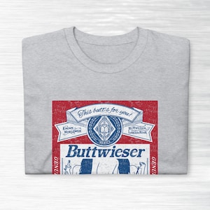 Peut inclure: Un t-shirt gris avec un graphique rouge, blanc et bleu d'une canette de bi&egrave;re parodique. La canette dit "Buttweiser" et "This butt's for you!"