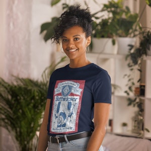 Peut inclure: Un t-shirt bleu marine avec un motif de label de bi&egrave;re Budweiser de style vintage. Le label a un fond rouge avec du texte et des images blancs et bleus. Le texte sur le label indique "Budweiser" et "Genuine Draft".
