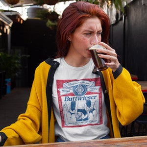 Peut inclure: Une femme portant un t-shirt blanc avec un graphisme d'&eacute;tiquette de bi&egrave;re de style vintage qui dit "Buttweiser" et un cardigan jaune. L'&eacute;tiquette pr&eacute;sente un fond rouge avec du texte bleu et des illustrations de deux femmes en bikini. Elle tient un verre de bi&egrave;re brune et le regarde.