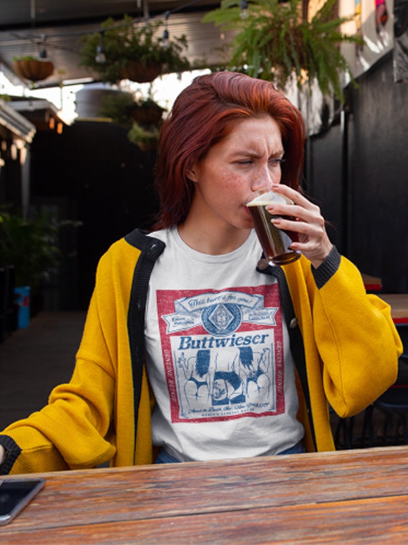 Peut inclure: Une femme portant un t-shirt blanc avec un graphisme d'&eacute;tiquette de bi&egrave;re de style vintage qui dit "Buttweiser" et un cardigan jaune. L'&eacute;tiquette pr&eacute;sente un fond rouge avec du texte bleu et des illustrations de deux femmes en bikini. Elle tient un verre de bi&egrave;re brune et le regarde.