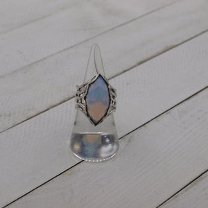 May include: A silver ring with a large, iridescent, light blue and pink gemstone. The ring is on a clear plastic ring display stand.