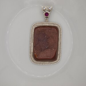 May include: A silver pendant with a brown agate stone set in a beaded frame. The pendant has a small pink gemstone at the top.