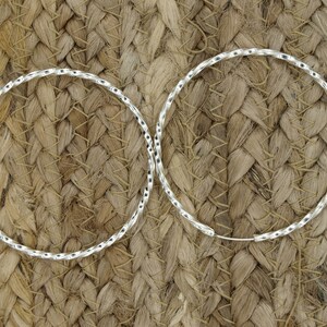 May include: A pair of silver hoop earrings with a twisted, textured design. The earrings are on a woven background.