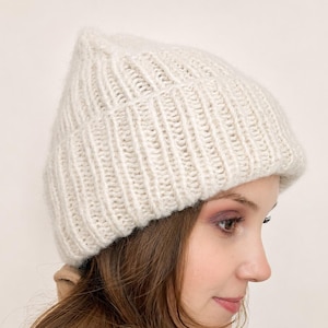 May include: A cream-colored knit beanie with a thick, vertical ribbed texture. The hat features a folded brim and a soft, fuzzy yarn construction, providing a structured yet cozy appearance that covers the top of the head down to the ears.